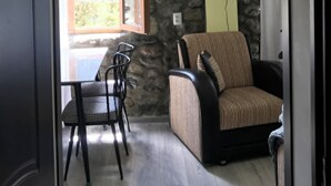 Un interior con pared de piedra, televisor sobre un sofá, mesa de comedor con sillas y una ventana con persiana abierta.
