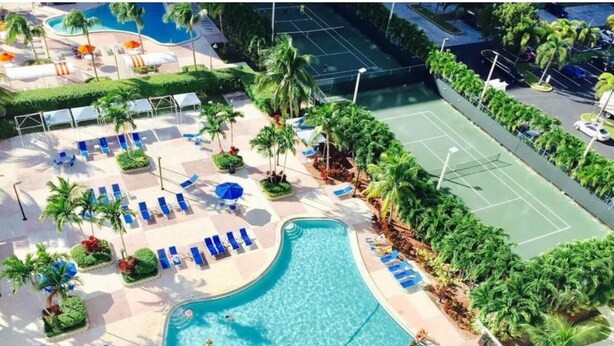 A swimming pool with lounge chairs, a tennis court, and palm trees. - Modern Sunny Isles Condo Near Beach (Sunny Isles Beach)