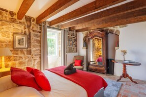 Ein Schlafzimmer mit Steinwand, Holzbalken, einem Bett mit roten Kissen, einem Spiegel und einem kleinen Tisch.