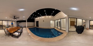 Ein moderner Poolbereich im Innenbereich mit Holztisch und -stühlen, Deckenventilator und einer gewölbten Glaswand.