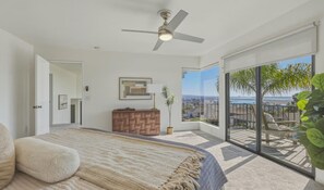Ein Schlafzimmer mit einem Bett, einem Deckenventilator, einem Nachttisch, einer Topfpflanze und Blick auf einen Balkon mit Geländer.