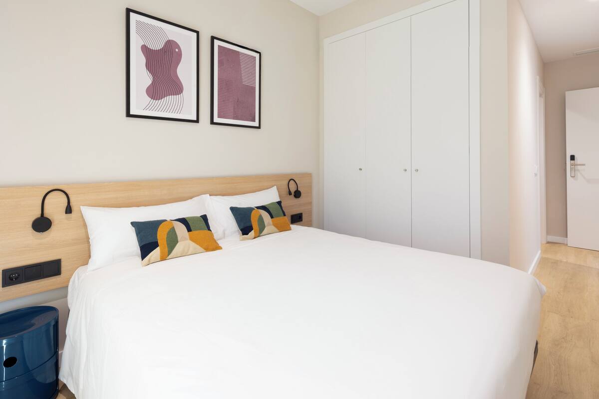 Un dormitorio con una cama, dos cuadros enmarcados, un armario y una mesita de noche con lámpara.