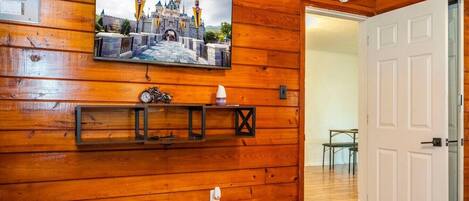 Interior de madera con un cuadro colgado de un castillo, una estantería con una motocicleta y una taza, y una puerta blanca.