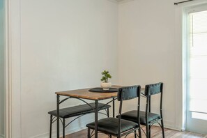 Ein Eckzimmer mit Holztisch, zwei schwarzen Stühlen, einem kleinen Topfpflanzen und einem Fenster mit Jalousien.