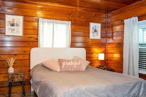 Una habitación de cabaña de madera con una cama, una mesita de noche, una lámpara y dos cuadros en la pared.