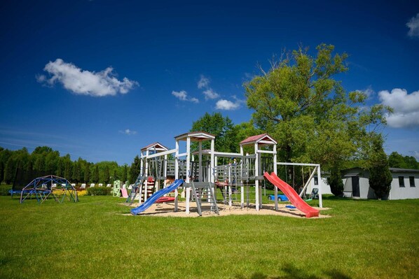 A playground with multiple play structures including slides and climbing areas, set in a grassy field with trees in the background.