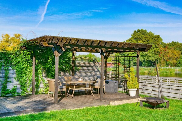 A wooden pergola with a table and chairs, surrounded by greenery and a body of water.