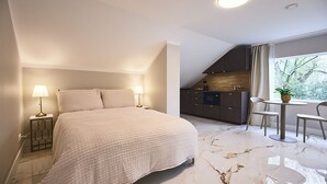 Ett sovrum med en säng, sänglampor, ett litet bord, en stol och utsikt över ett kök och en balkong.