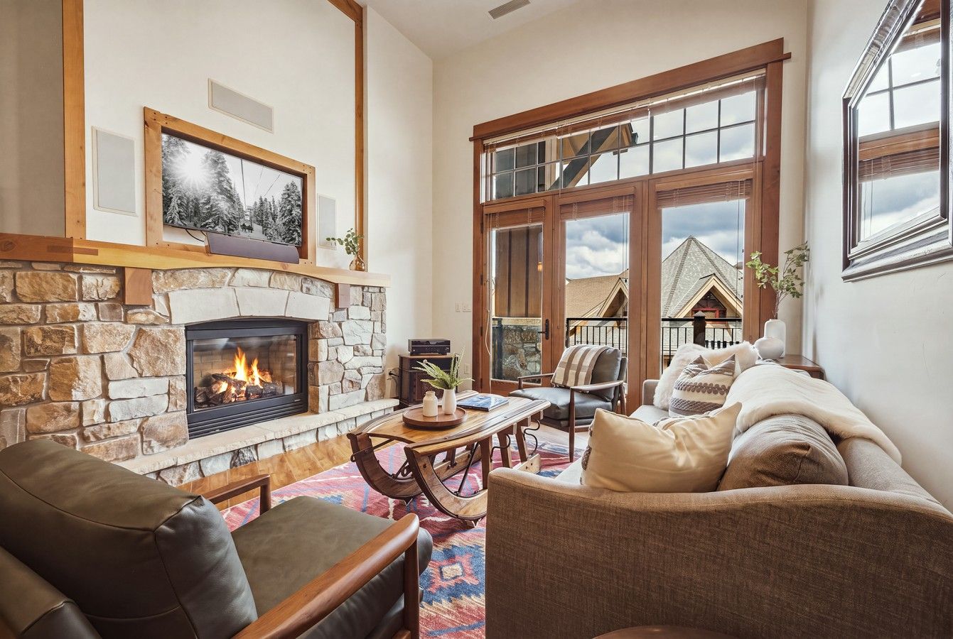 A cozy living room with a stone fireplace, leather sofas, a coffee table, and a large window with a view of a house.