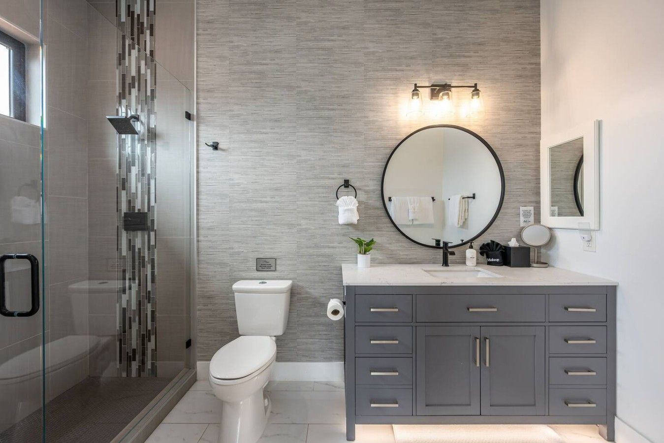 A modern bathroom with a glass shower enclosure, a toilet, a vanity with a mirror, and a wall-mounted light fixture.