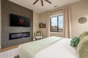 Habitación de hotel con cama, televisión montada en la pared, chimenea, ventana con cortinas y una silla.