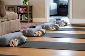 A yoga mat setup with a grey cushion and a rolled-up blanket on a wooden floor, a grey sofa, and a bookshelf in the background.