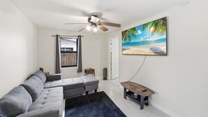 A living room with a grey sofa, a ceiling fan, a painting of a beach, and a small wooden table. - Delightful 3-bedroom house with AC in West Palm Beach (West Palm Beach)