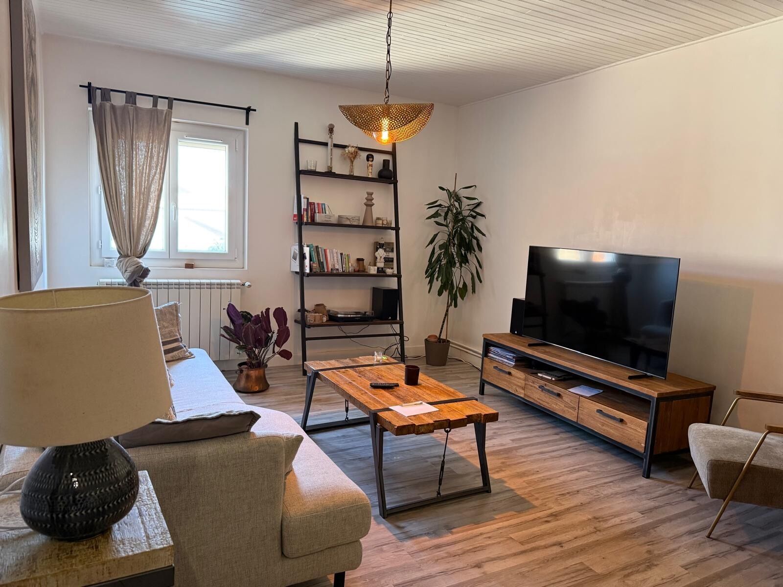 En moderne stue med sofa, sofabord, fjernsyn, reol og en hængende lampe.
