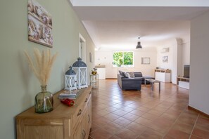 Een ruime woonkamer met een bank, een bijzettafeltje en een eettafel. Er is een raam met uitzicht op groen, en een decoratieve vaas op een houten kast.