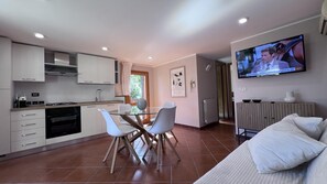 Eine moderne Küche mit weißen Schränken, einem Esstisch mit Stühlen, einem Fernseher an der Wand und einer Wohnbereich mit Sofa und Heizkörper.