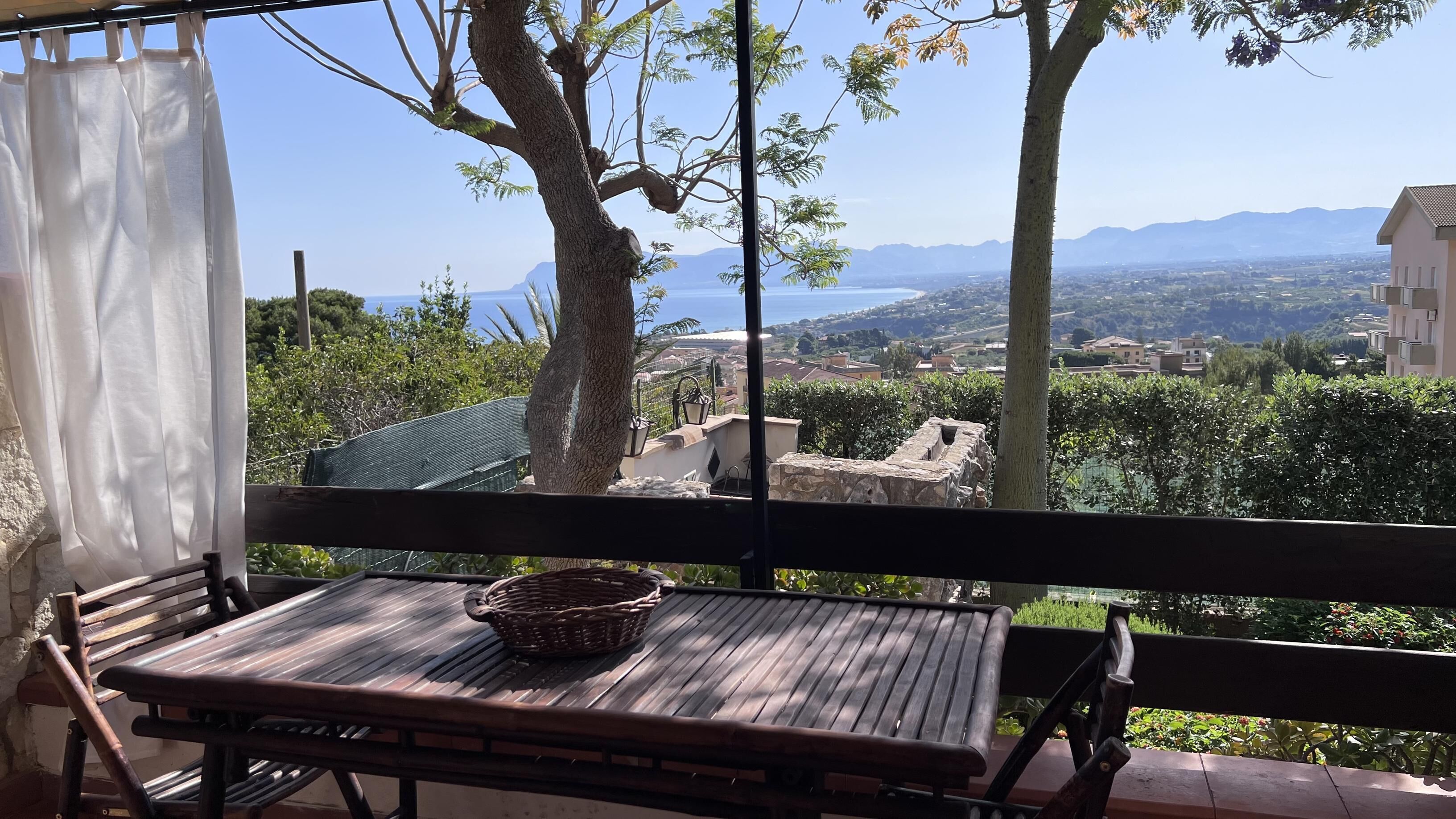 Un balcon avec une table et des chaises en bois, un panier en osier et une vue sur un paysage côtier avec des montagnes et des bâtiments.