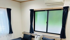 Ein Zimmer mit zwei Betten, einem Fenster mit Vorhängen, einer Klimaanlage und einem kleinen Tisch.