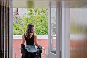 Eine Person steht an der Balkontür und blickt auf einen Garten mit roter Wand und viel Grün.