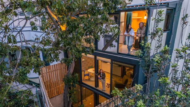 Ett modernt hus i två våningar med en glasfasad, ett trädäck och en inredning som syns utifrån, inklusive ett matområde.