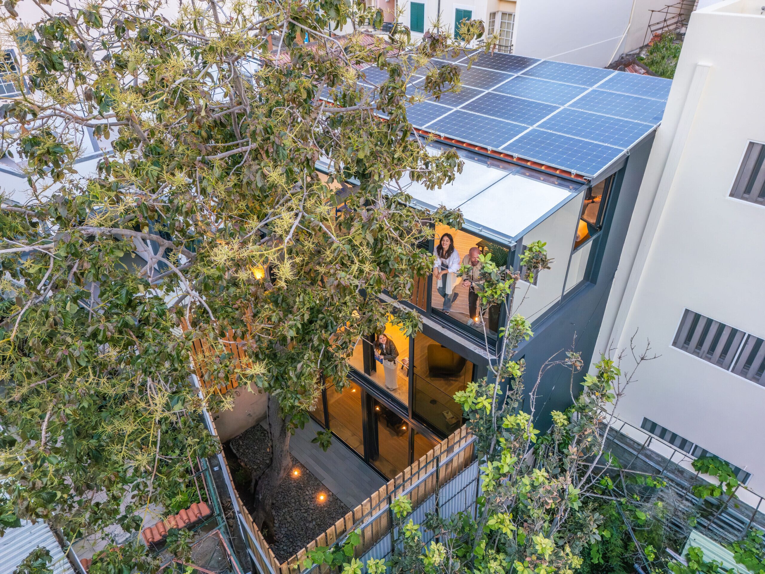 Um edifício de vários andares com um painel solar no terraço, uma varanda com vista para a rua e uma árvore em primeiro plano.