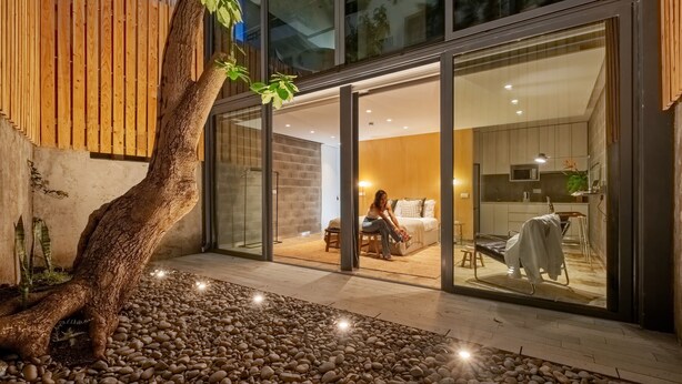 Ein modernes Interieur mit einem großen Glasfenster, das in ein Schlafzimmer führt, einem sichtbaren Küchenbereich und einem Baum im Vordergrund.