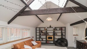 Une pièce de style « loft », avec un plafond haut, des poutres apparentes, une grande fenêtre avec stores, un canapé en cuir marron, un plaid blanc, une bibliothèque, un fauteuil en forme de sac de haricots noir et un bureau avec une lampe.