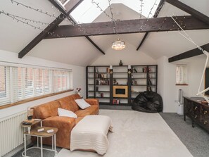 A loft-style room with a high ceiling, exposed beams, a large window with blinds, a brown leather sofa, a white throw blanket, a bookshelf, a black bean bag, and a desk with a lamp. - The Joinery (Scarborough)