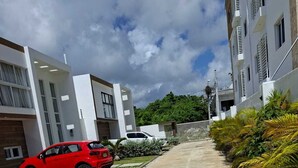A modern residential street with a red car parked on the left, a concrete pathway, and a row of palm trees on the right. - 3-bedroom house in charming Punta Cana with cool AC (Punta Cana)