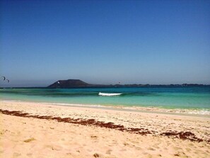 Een strand met helder turquoise water, een zandstrand en een eiland in de verte.
