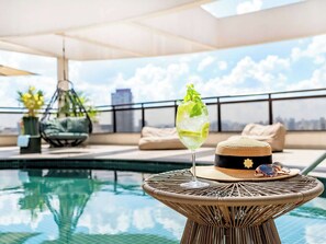Ein Pool auf dem Dach mit einem Glas grünes Getränk, einem Korbstuhl und Blick auf die Stadt.