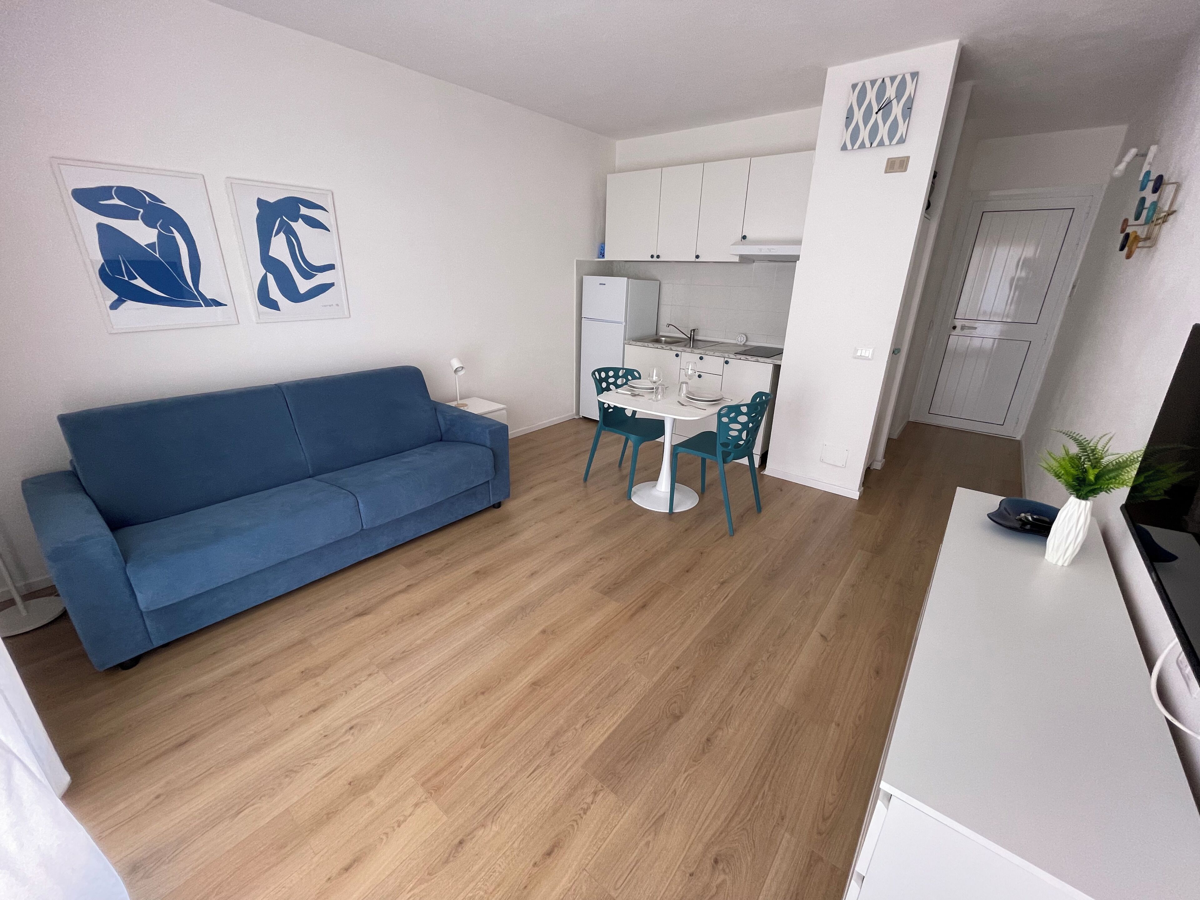Una sala de estar moderna con un sofá azul, un comedor con una mesa blanca y sillas color turquesa, y una cocina con armarios blancos.