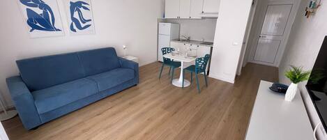 Una sala de estar moderna con un sofá azul, un comedor con una mesa blanca y sillas color turquesa, y una cocina con armarios blancos.