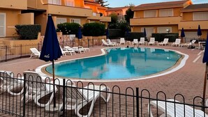 Ein Swimmingpool, umgeben von Liegestühlen und Sonnenschirmen, mit einem Zaun im Vordergrund.