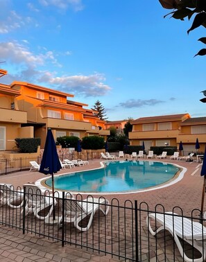 A swimming pool surrounded by lounge chairs and umbrellas, with a fence in the foreground. - Studio apartment in the center of Badesi with shared pool (Badesi, provincia di Sassari)