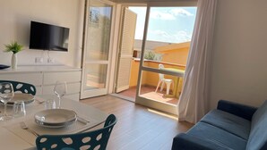 Ein Zimmer mit Esstisch für zwei Personen, einer Couch, einem an der Wand befestigten Fernseher und einer Glastür zum Balkon mit Tisch und Stühlen.