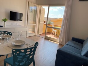 A room with a dining table set for two, a sofa, a TV mounted on the wall, and a sliding glass door leading to an outdoor area with a table and chairs. - Studio apartment in the center of Badesi with shared pool (Badesi, provincia di Sassari)