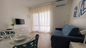 Ein modernes Wohnzimmer mit einer Couch, einem Esstisch für zwei Personen, einem Fernseher und einem Fenster mit Vorhängen.