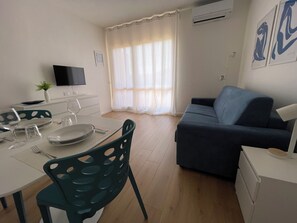 A modern living room with a sofa, a dining table set for two, a TV, and a window with curtains. - Studio apartment in the center of Badesi with shared pool (Badesi, provincia di Sassari)