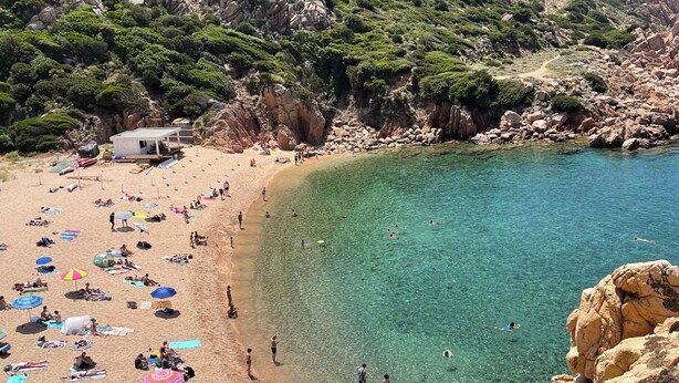 Ein Strand mit klarem Wasser, felsigem Gelände und einem kleinen Gebäude.