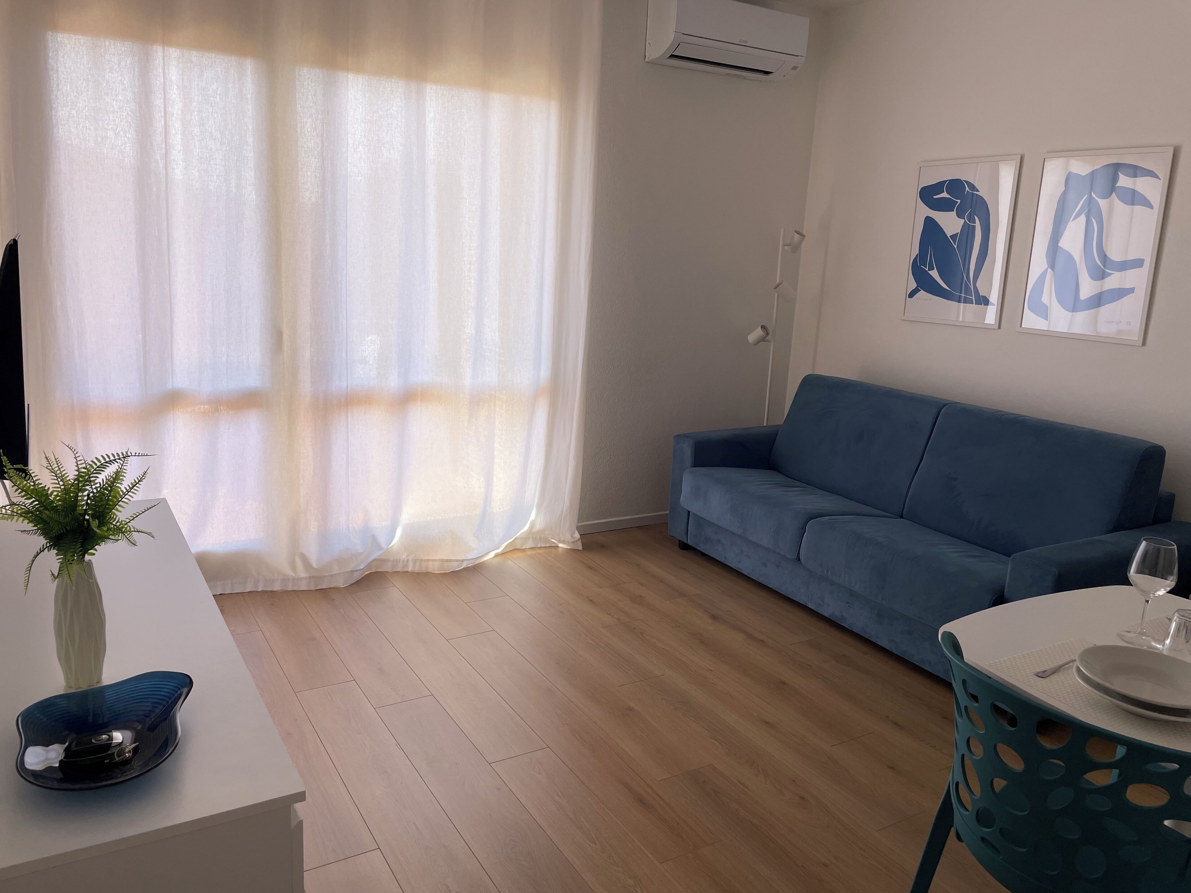 Un salón con un sofá azul, una mesa blanca con un vaso y una silla azul.