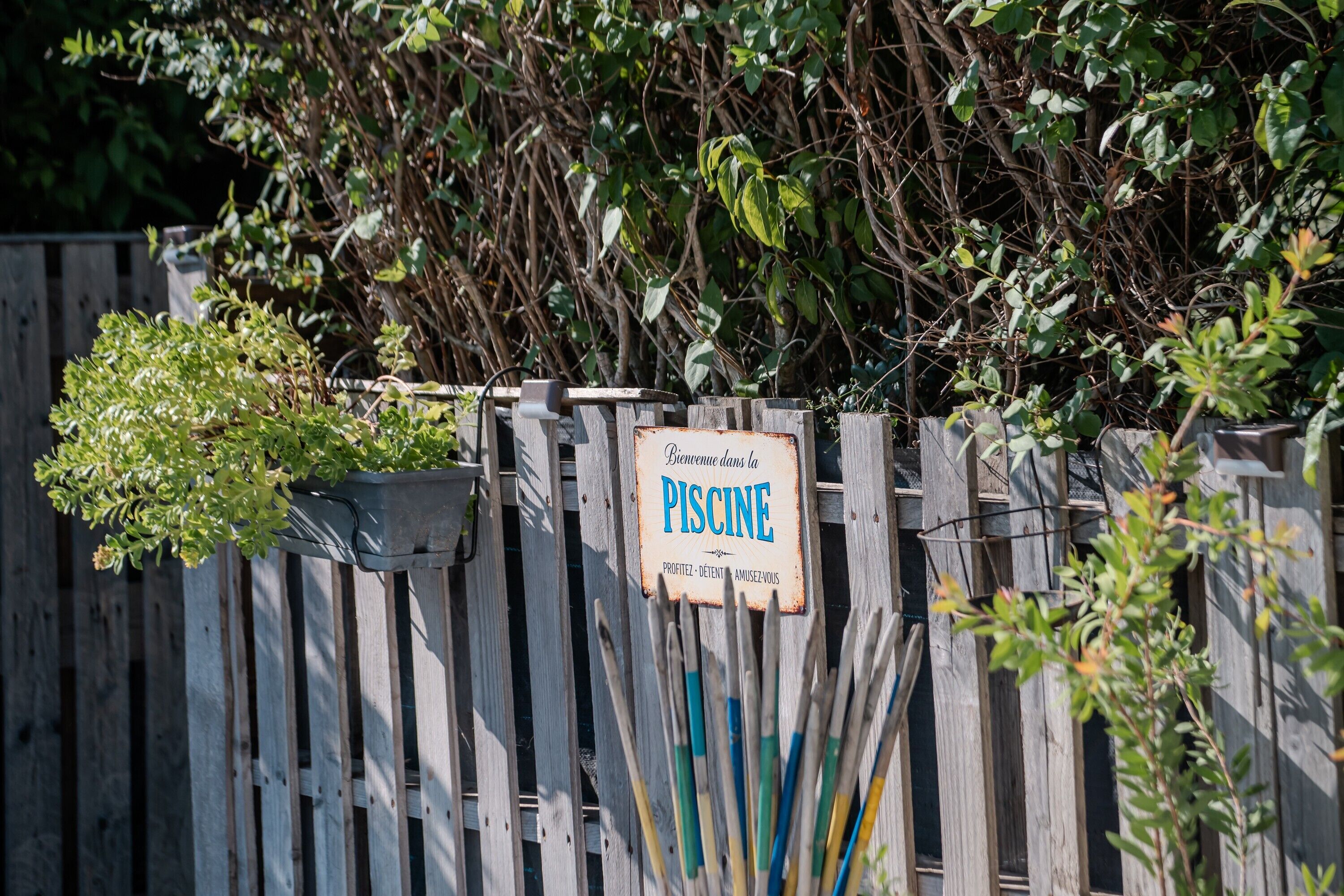 Une clôture en bois avec une pancarte sur laquelle on peut lire « Bienvenue à la PISCINE » et un pot de fleurs placé sur la gauche.
