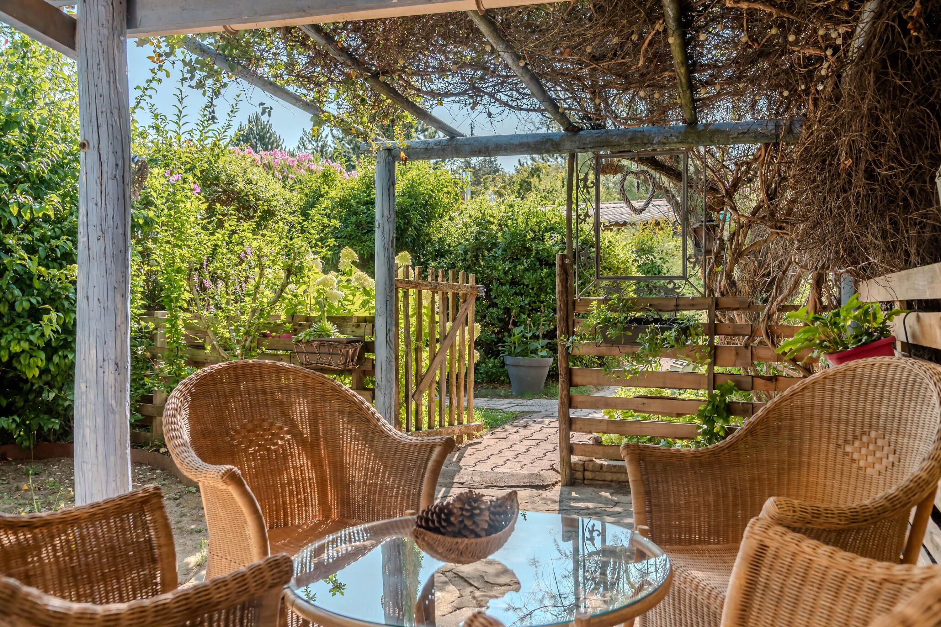 A covered outdoor seating area with wicker chairs and a glass table, surrounded by lush greenery and a wooden fence.