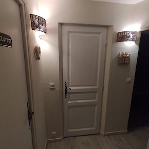 Un pasillo con una puerta blanca, un aplique en la pared y un estante pequeño con objetos al lado derecho.