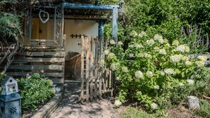Una estructura de madera rústica con una puerta y un pequeño porche, rodeada de abundante follaje y plantas en flor.