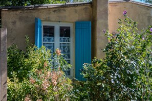A rustic building with a window and blue shutters, surrounded by lush greenery. - “Tamaris Les Floralies in Provence” with Mountain View, Shared Pool and Private Terrace (Malaucène)