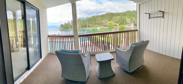 A balcony with wicker chairs and a small table, overlooking a lake with houses and trees.