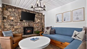 Ein gemütliches Wohnzimmer mit Kamin aus Stein, einem blauen Sofa, einem braunen Ledersessel und einem runden weißen Tisch.