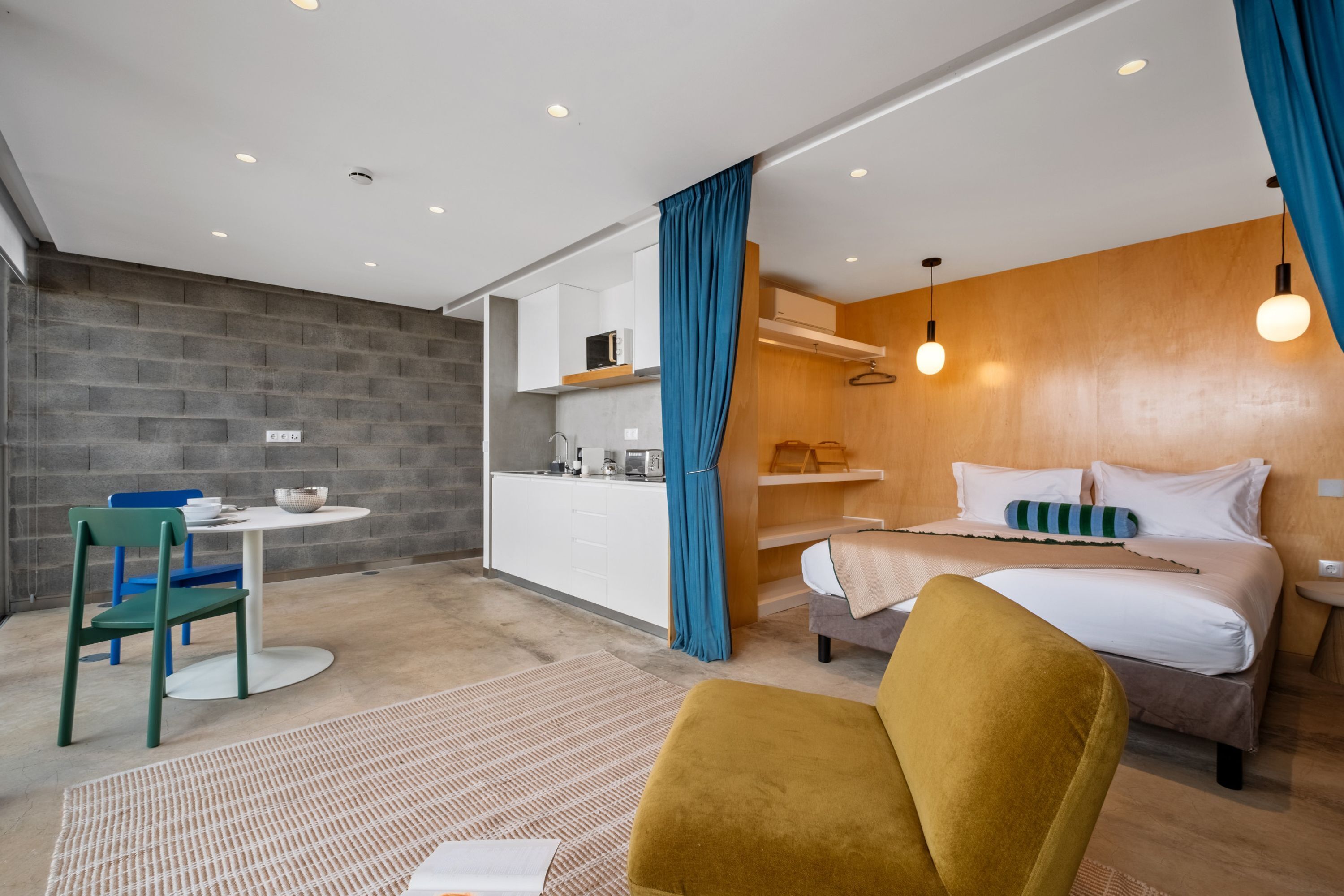 Une chambre d’hôtel moderne équipée d’une kitchenette, d’un lit avec un oreiller à rayures, d’un rideau bleu et d’un fauteuil de couleur moutarde.