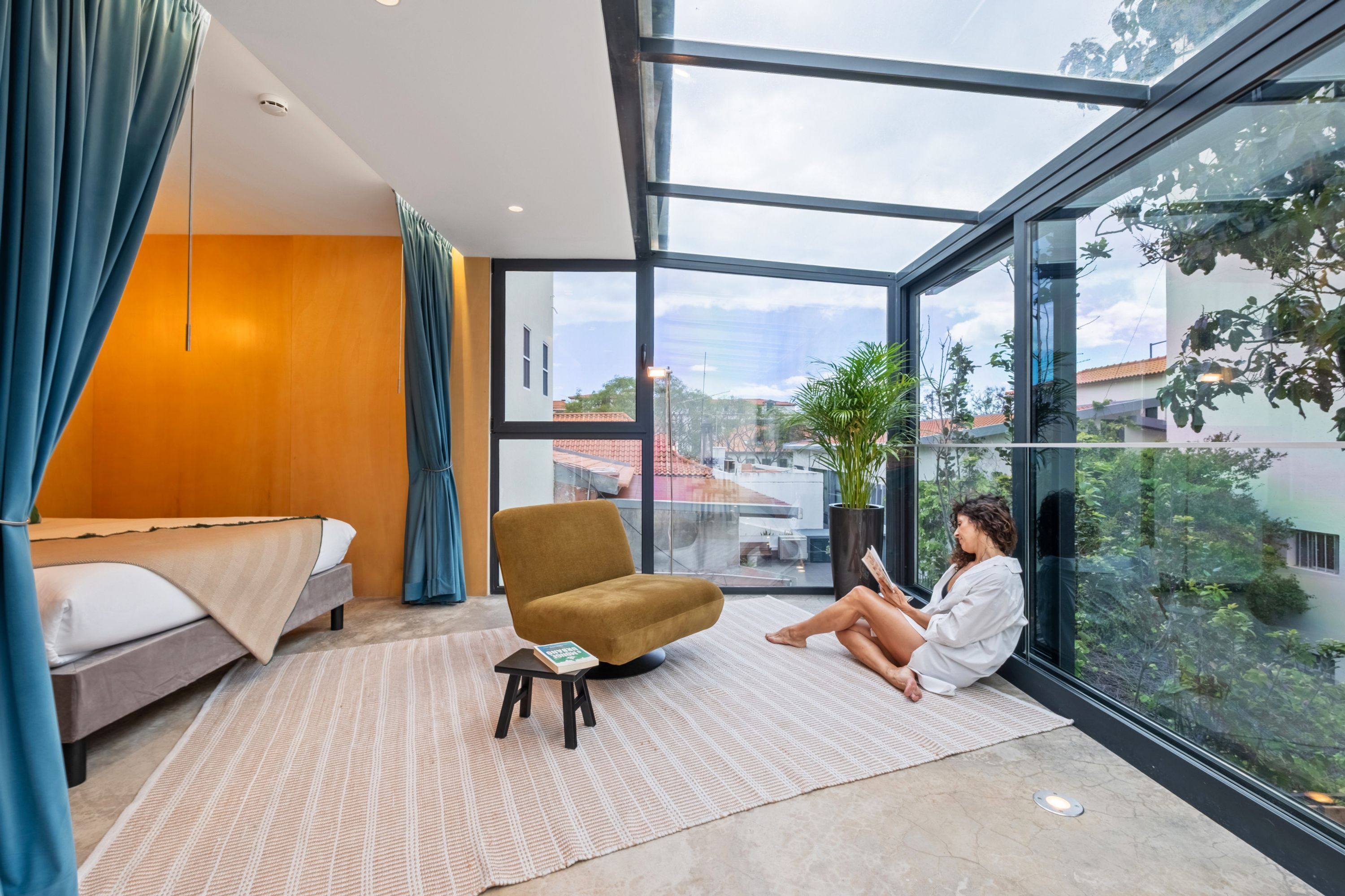 Una habitación de hotel moderna con un ventanal grande, una cama, un sillón de lectura y una pequeña mesa con un libro.